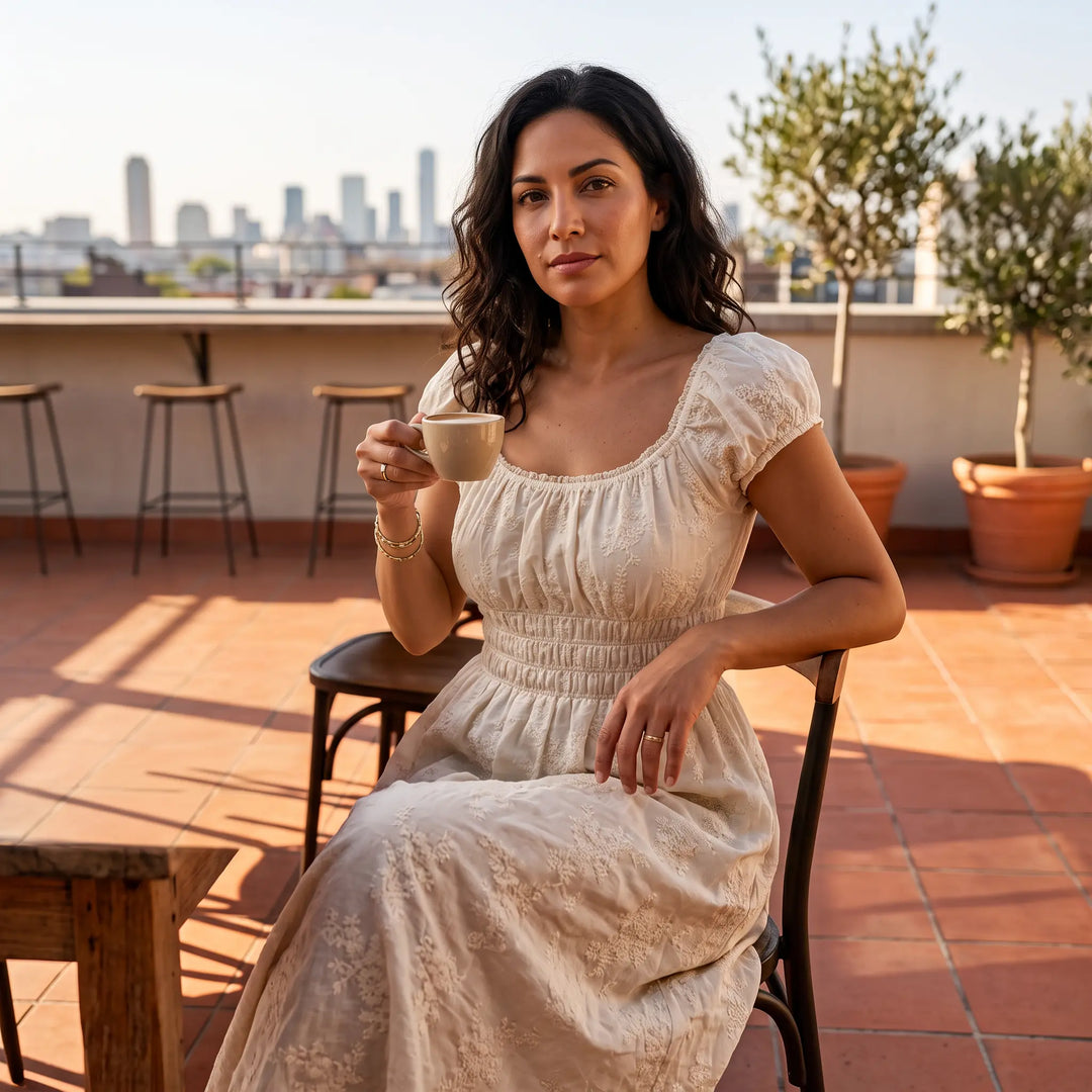 A woman enjoys a coffee while wearing the Altera soft ivory embroidered midi dress featuring a square neckline, puff sleeves, and a smocked bodice, seated on an outdoor terrace.