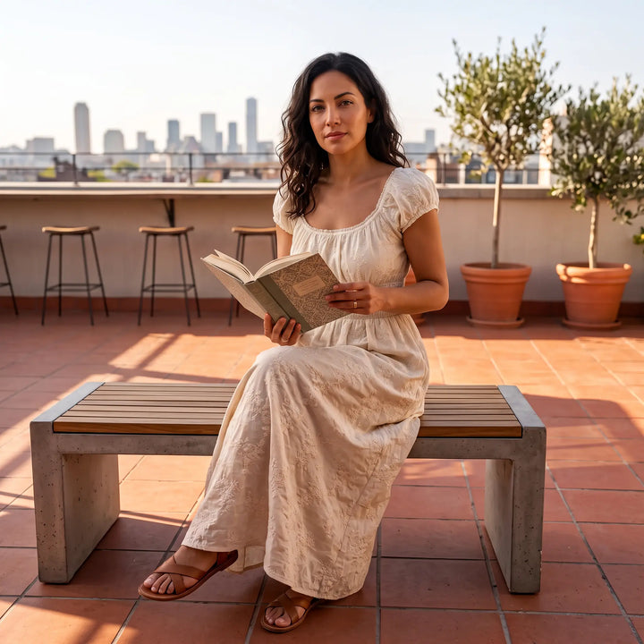 A woman wears a flowing beige embroidered midi dress with a square neckline and puff sleeves while reading a book on an outdoor bench with a cityscape in the background.