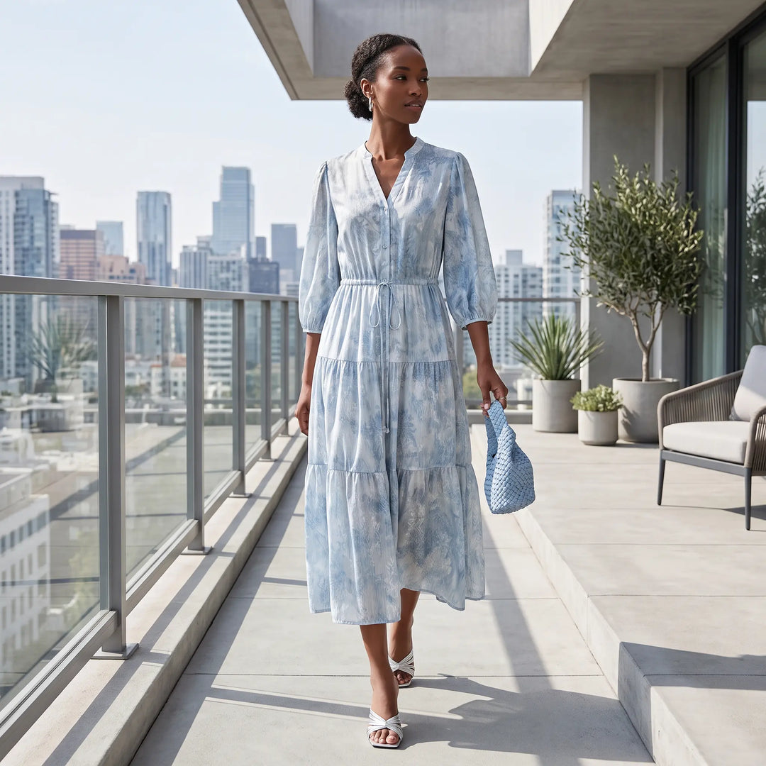 Woman walking on a city balcony wearing a flowing sky blue and white watercolor print silk blend midi dress with tiered skirt and balloon sleeves.