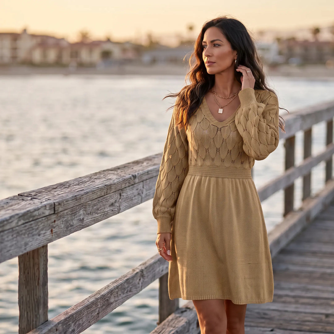 A woman wearing a rich camel V-neck midi knit dress with a fish-scale pattern on the yoke and sleeves stands on a wooden pier by the water.