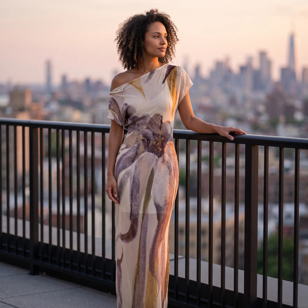 Woman wearing an elegant Kaelen flowing silk blend maxi dress with an abstract floral print in muted purples and cream, standing on a rooftop with a city skyline at dusk.