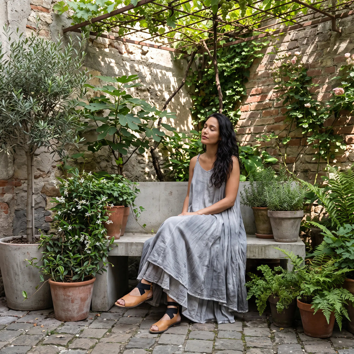 A woman relaxes in a serene urban garden wearing a flowing, plant-dyed grey cotton maxi dress with an irregular hem and delicate spaghetti straps.
