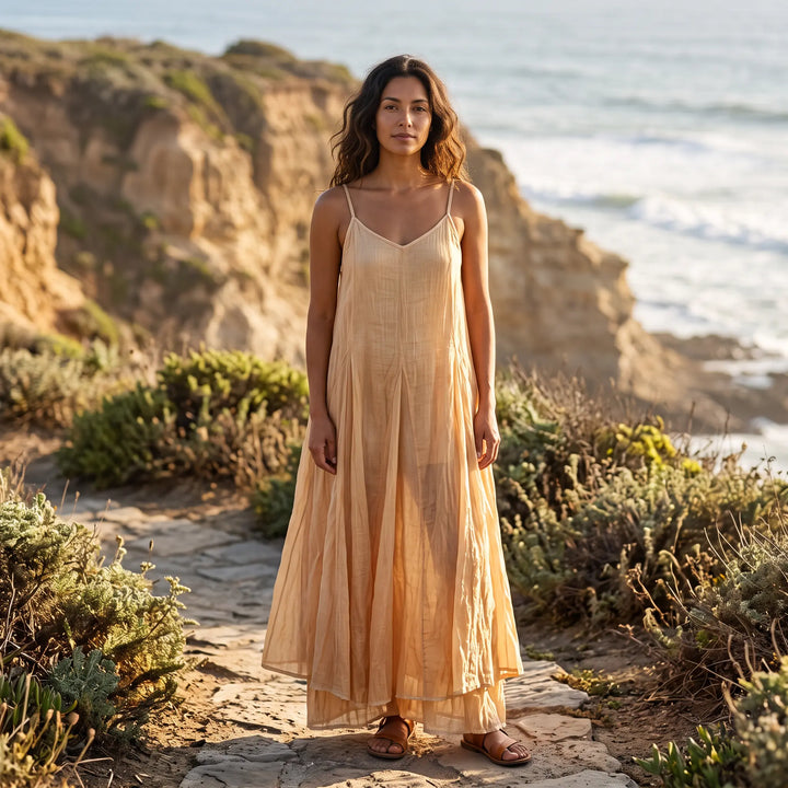 A woman models a flowing, plant-dyed cotton maxi dress with adjustable spaghetti straps and a V-neckline, standing on a stone path by the ocean. The dress is a light peach hue with subtle variations and features an irregular hem.