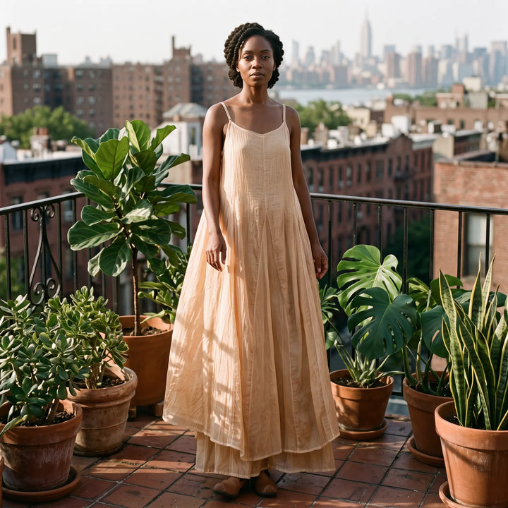 A woman models a flowing, loose-fitting Kaelen plant-dyed cotton maxi dress in a soft, peachy-beige hue on a city balcony surrounded by lush plants.