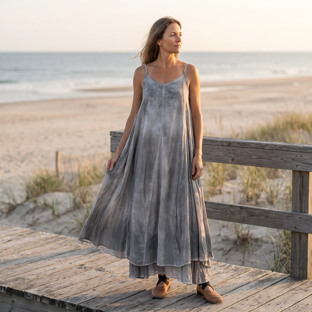 A woman stands on a wooden boardwalk by the ocean, wearing a flowing, cool grey plant-dyed cotton maxi dress with adjustable spaghetti straps and an irregular hemline.