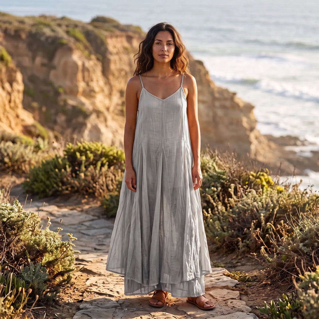 Female model wearing a Kaelen sustainable plant-dyed cotton maxi dress in Cool Grey Mist, featuring a V-neck, spaghetti straps, and an irregular flowing hem, standing outdoors by the coast.