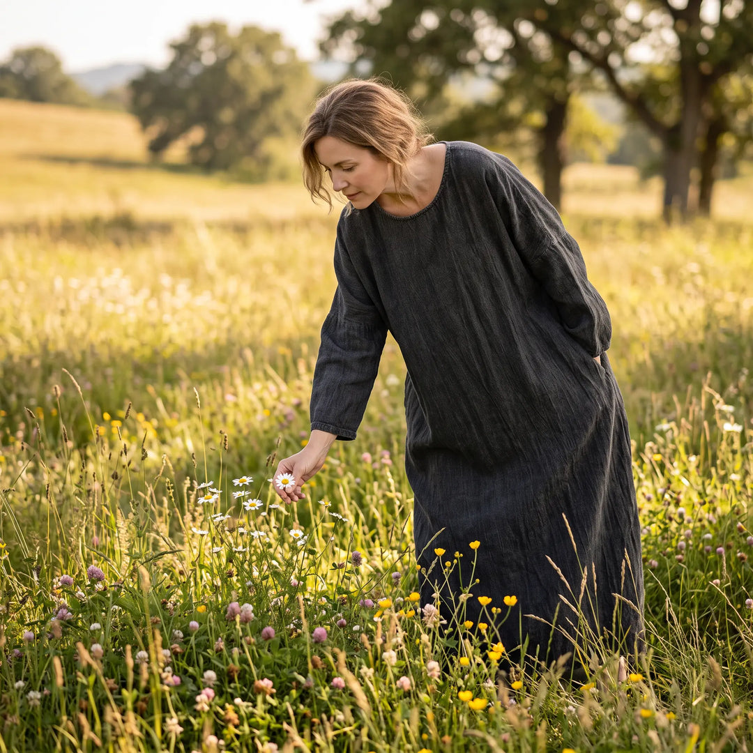 Woman wearing a charcoal grey Kaelos garment-washed cotton-linen maxi dress, reaching for a daisy in a sunlit meadow.