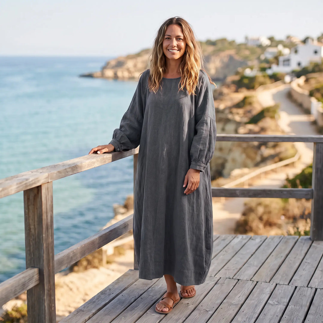 Woman wearing a charcoal grey Kaelos oversized linen blend maxi dress with wide sleeves and a gathered round neck, standing on a wooden deck by the sea.