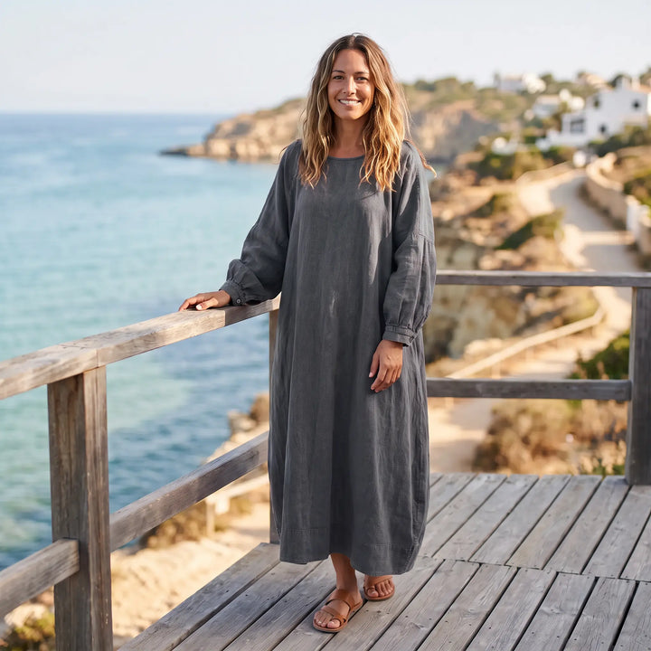 Woman wearing a charcoal grey Kaelos oversized linen blend maxi dress with wide sleeves and a gathered round neck, standing on a wooden deck by the sea.