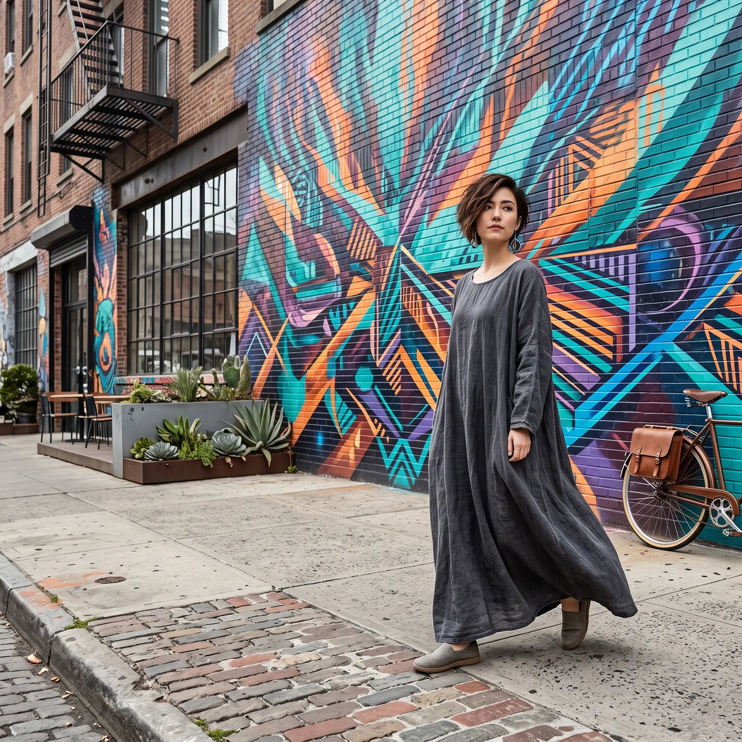 A woman walks along a city street wearing a flowing charcoal grey Kaelos linen blend maxi dress, with a vibrant mural in the background.