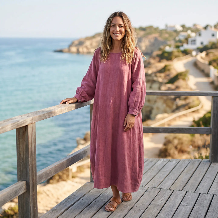 A woman smiles, wearing a flowing muted rose cotton-linen blend maxi dress with wide sleeves, standing on a wooden deck overlooking the sea.