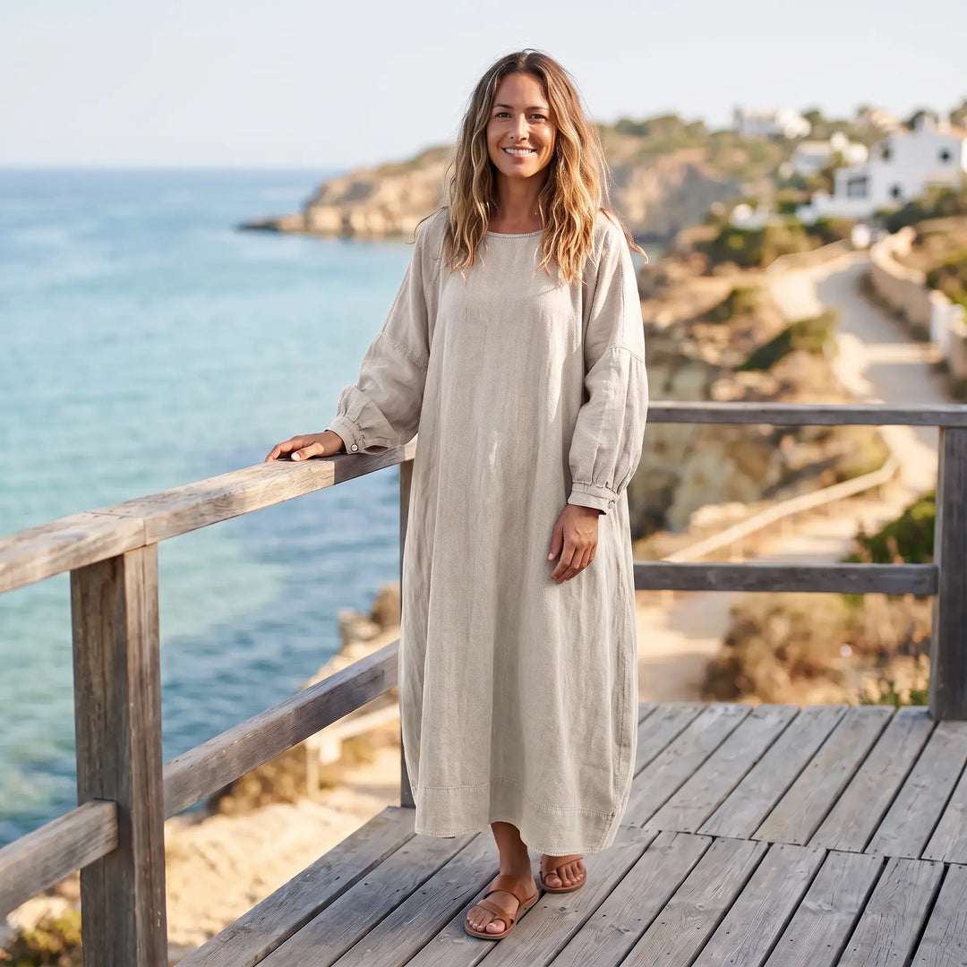 A woman stands on a wooden deck overlooking the ocean, wearing a long, loose-fit oat beige cotton-linen blend dress with wide sleeves and side slits.