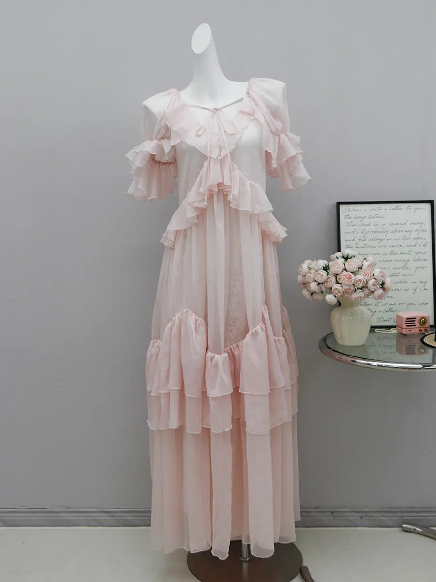 Pale blush pink Kynari maxi dress featuring tiered ruffle detailing, sheer georgette overlay with an embossed lace pattern, and adjustable V-neckline with tie closure, displayed on a mannequin.