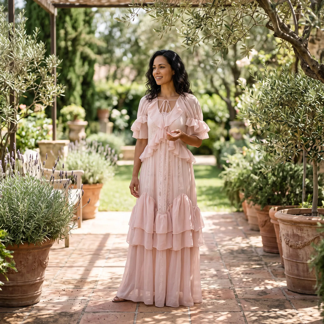 A woman models an ethereal pale blush pink tiered ruffle maxi dress with sheer georgette overlay and flutter sleeves while walking in a sun-dappled garden.