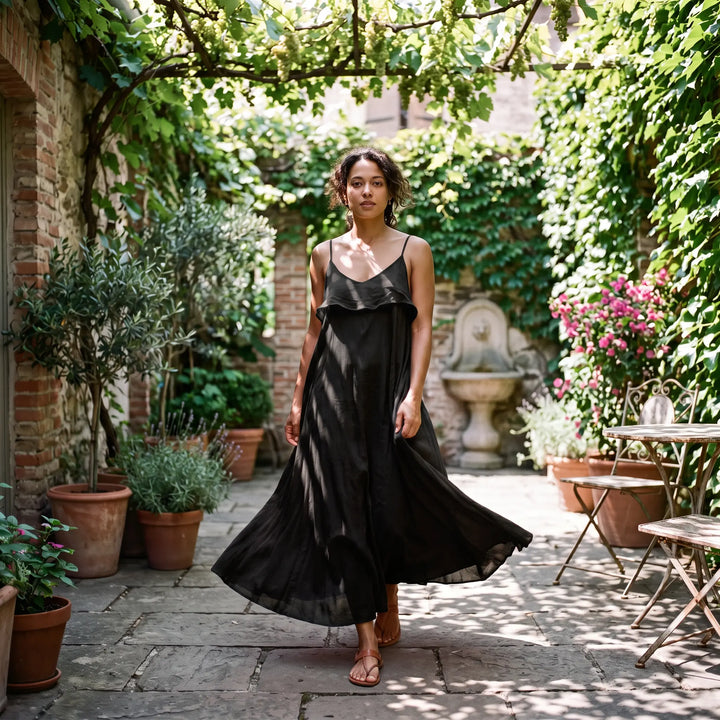A woman in a flowing black pure cotton maxi dress with thin spaghetti straps and a ruffled V-neckline walks outdoors, her voluminous skirt twirling.