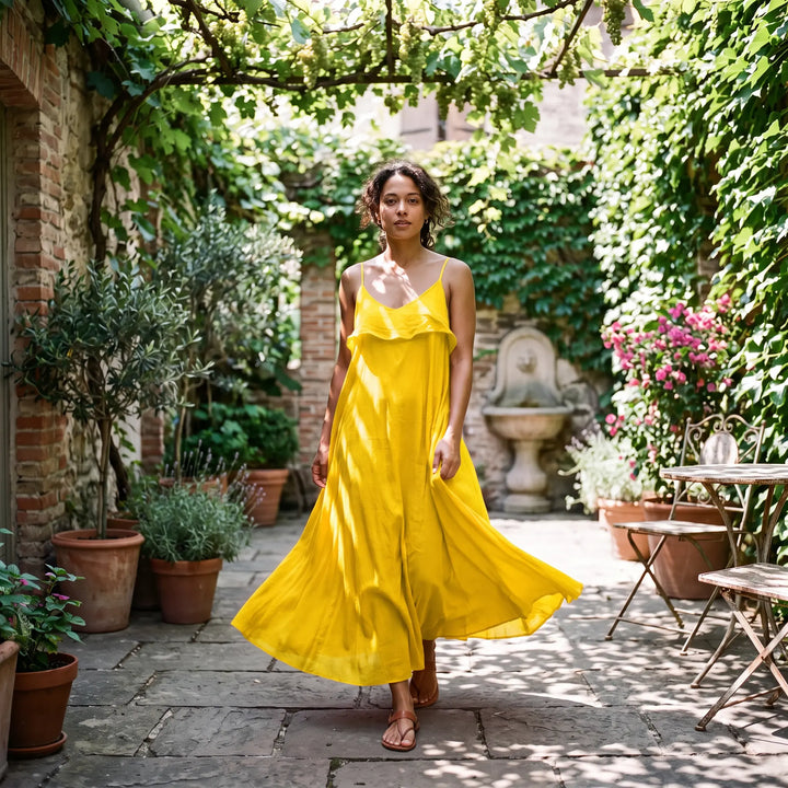 A woman walks through a sun-dappled garden wearing a vibrant sunny yellow pure cotton maxi dress with thin spaghetti straps and a flowing A-line skirt featuring a V-neckline with raw-edged ruffles.