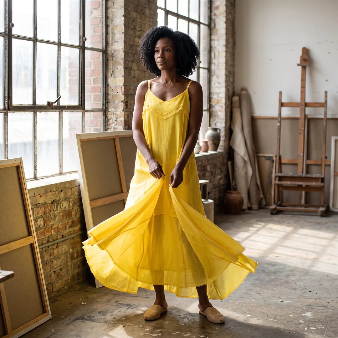 A woman models a flowing, sunny yellow pure cotton maxi dress with thin spaghetti straps and a V-neckline in a bright artist studio. The voluminous skirt is caught in motion, showcasing its raw-edged hem.