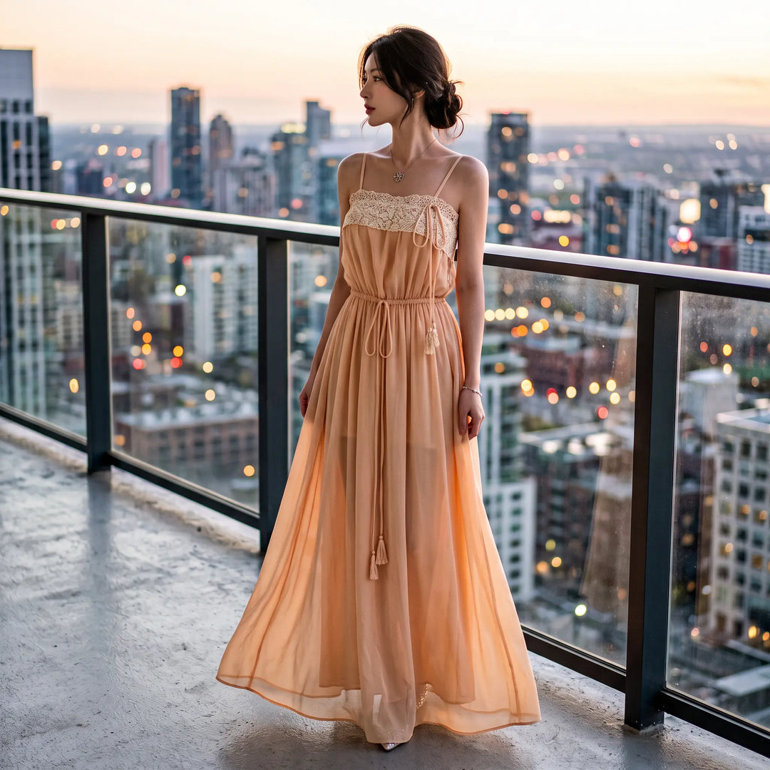 Woman wearing a pale peach-nude sheer chiffon maxi dress with a delicate floral lace bodice and spaghetti straps, standing on a balcony overlooking a city at sunset.