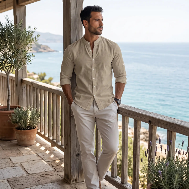 Man wearing a light beige long-sleeve linen-blend shirt with a Mandarin collar and light trousers, standing on a balcony overlooking the sea.