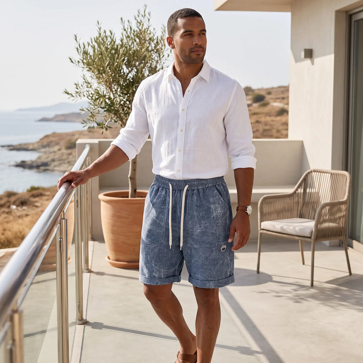 Man wearing blue textured mid-thigh shorts and a white linen shirt standing on a sunny balcony overlooking the sea, showcasing a relaxed summer style.