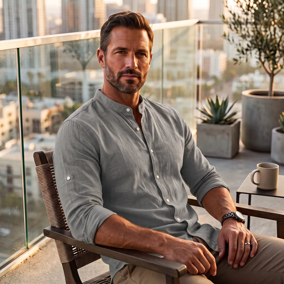 Man wearing a subtle iron grey long-sleeve linen-blend shirt with a Mandarin collar, seated on a balcony with a city view.