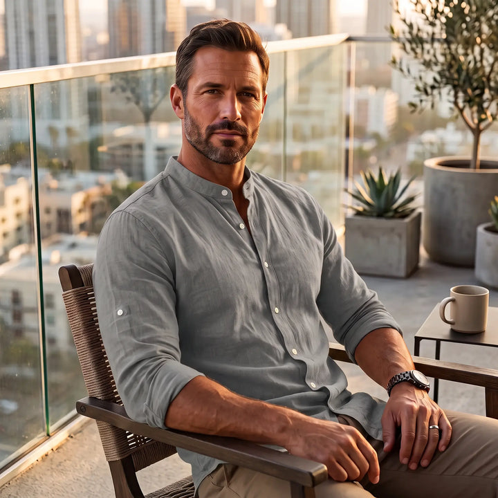Man wearing a subtle iron grey long-sleeve linen-blend shirt with a Mandarin collar, seated on a balcony with a city view.