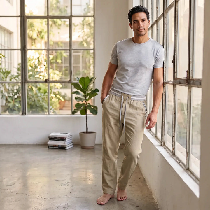 Man wearing warm khaki linen blend straight-leg trousers with a striped elastic waistband and drawstring, posing by a window in a sunlit room.