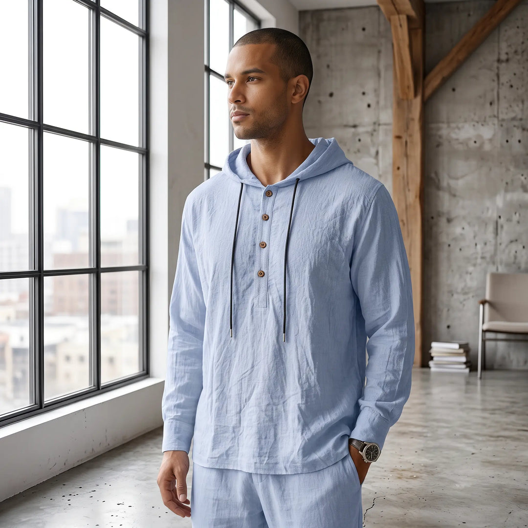 Man wearing a light blue linen blend loungewear set featuring a hooded henley top with a drawstring and trousers, in a relaxed fit, standing by a large window.