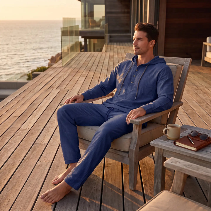Man relaxing on a wooden deck overlooking the ocean, wearing a navy blue hooded henley loungewear set made from a linen blend.