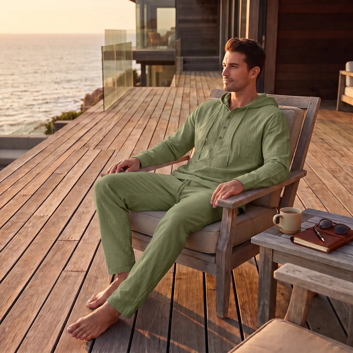 Man in an olive green linen blend loungewear set, featuring a hooded henley top and relaxed trousers, sitting on a wooden deck overlooking the ocean at sunset.