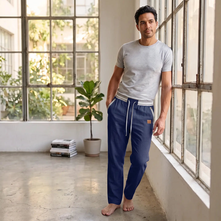 Man wearing deep navy Zylath linen blend trousers with an elasticated striped waistband and drawstring, leaning against a window. These relaxed-fit pants feature contrast pocket trim. Suitable for casual wear.