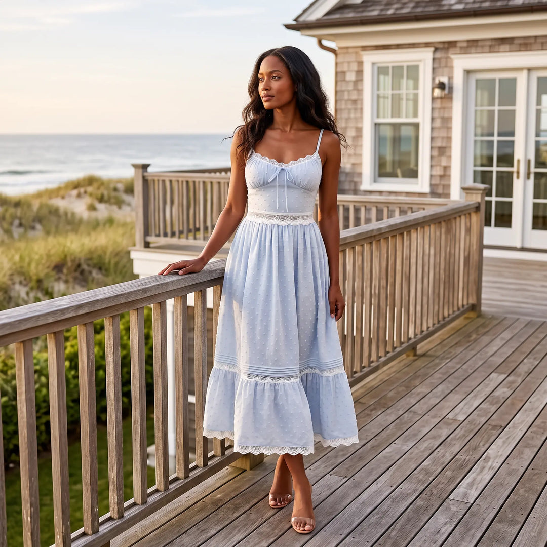 Pale blue polka dot lace maxi dress with spaghetti straps and a tie-front bodice, worn by a model on a seaside wooden deck with ocean and dunes in the background.