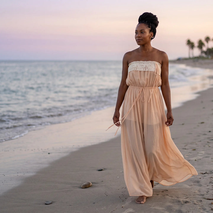 Woman walking on a beach at sunset, wearing a flowing pale peach-nude sheer chiffon maxi dress with a delicate lace trim bodice and an adjustable drawstring waist.
