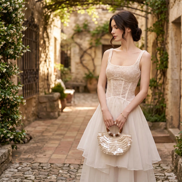 A sophisticated white sleeveless A-line dress with an intricate floral lace bodice and square neckline, paired with a flowing sheer midi skirt, modeled in a sunlit European-style courtyard.