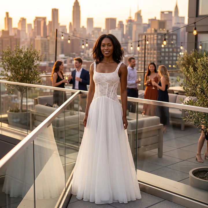 Woman smiling on a rooftop terrace at golden hour, wearing an elegant white sleeveless A-line midi dress with a detailed lace bodice and a flowing sheer skirt.