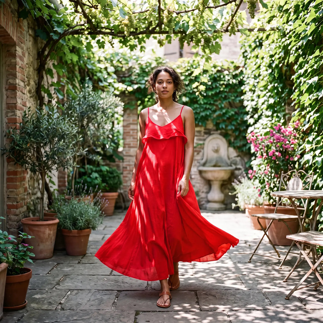 Woman walking in a vibrant red pure cotton maxi dress with spaghetti straps and a flowing A-line skirt, styled in an outdoor courtyard with lush greenery and stone pathways.