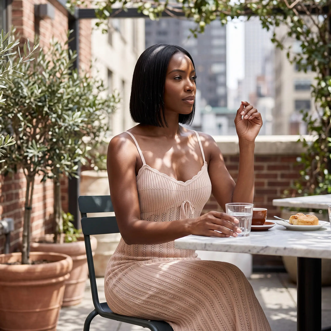 A woman in a soft Mist Twilight Blue ribbed knit midi dress with spaghetti straps and a drawstring V-neckline sits at a table on an outdoor terrace, enjoying a drink.