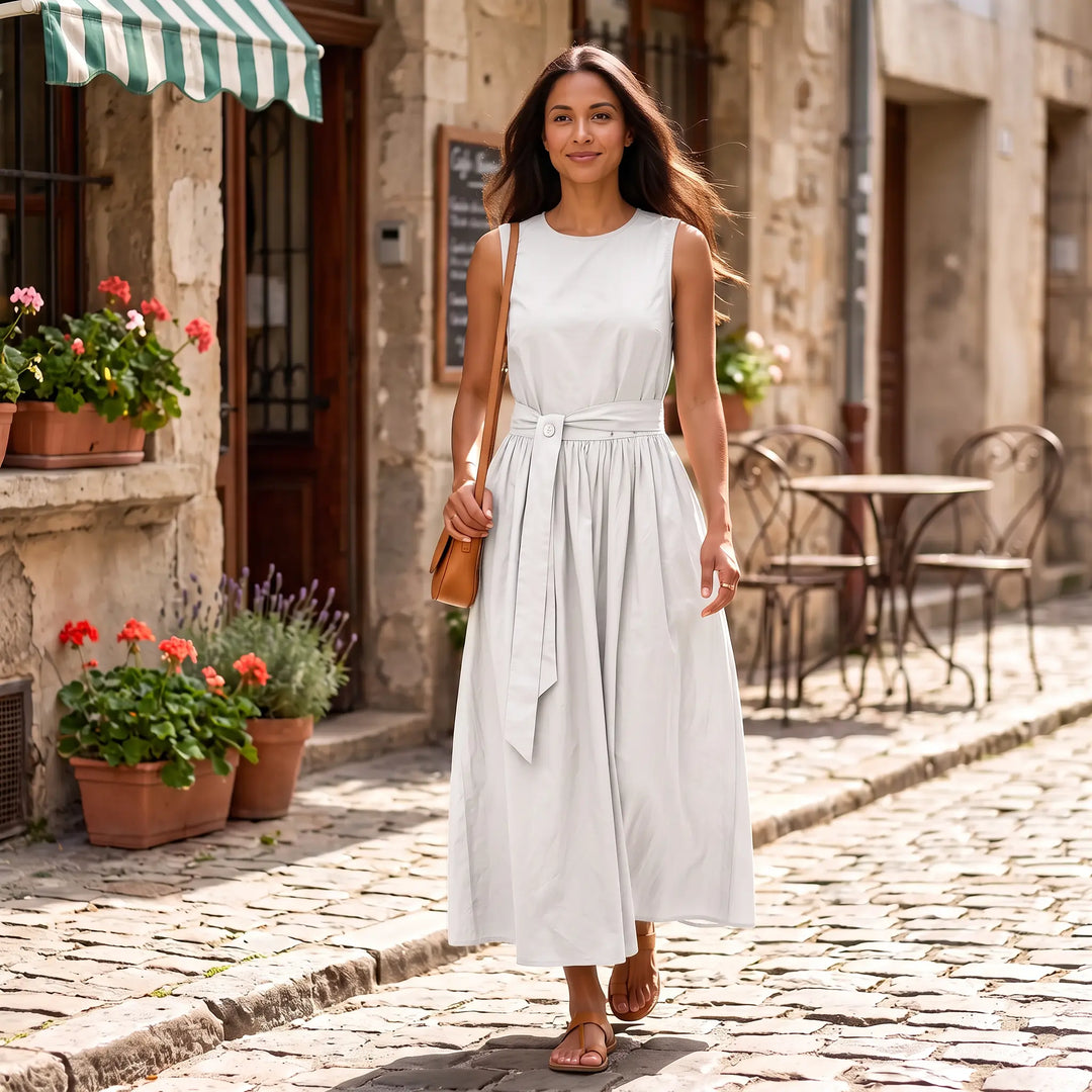 Woman walking down a charming European cobblestone street, wearing a sleeveless, off-white maxi dress with a cinched belted waist and flowing A-line skirt.