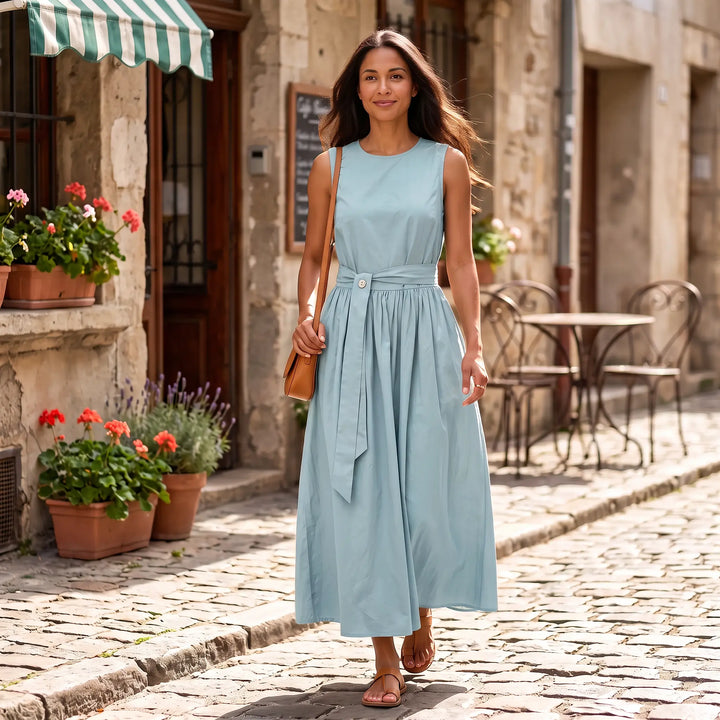 A woman walks down a cobblestone street wearing a sleeveless sky blue maxi dress with a cinched waist belt and a brown crossbody bag, showcasing its elegant flow and casual chic style.