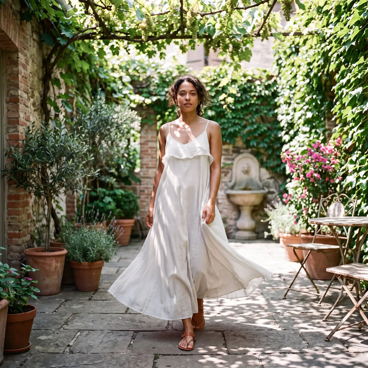 A woman walks towards the camera in a flowing pure white cotton maxi dress with thin spaghetti straps and a V-neckline, set in a charming Mediterranean courtyard.