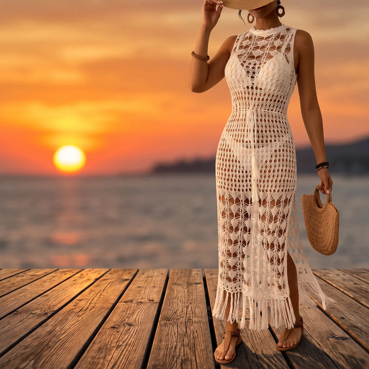 Woman wearing a white sleeveless crochet maxi dress with mock neckline, drawstring waist, high side slits, and fringe hem, posing on a wooden pier at sunset.