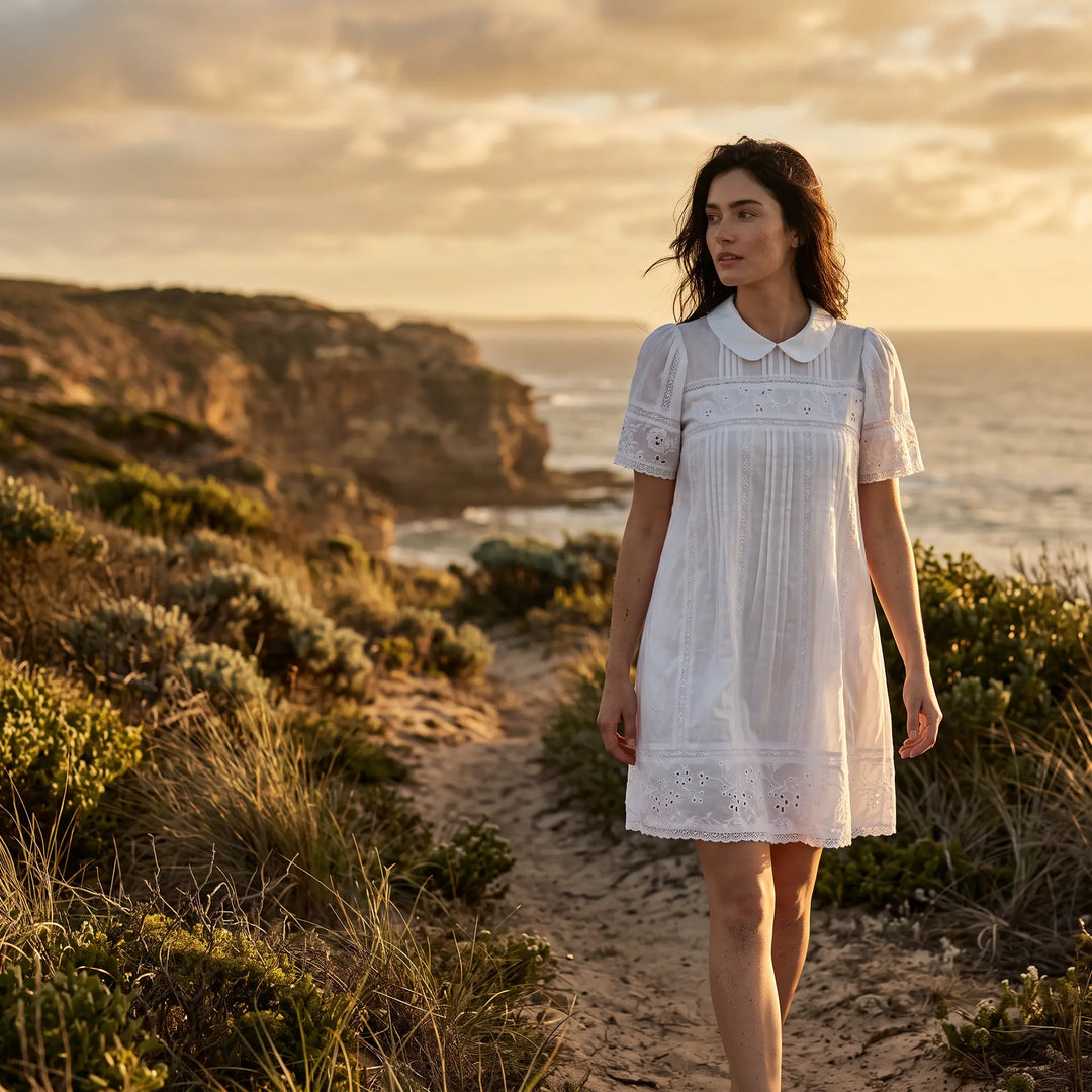 A woman models a white knee-length A-line cotton-blend dress, featuring a Peter Pan collar, puff sleeves, intricate embroidery, and lace trim, walking on a scenic coastal path at sunset.