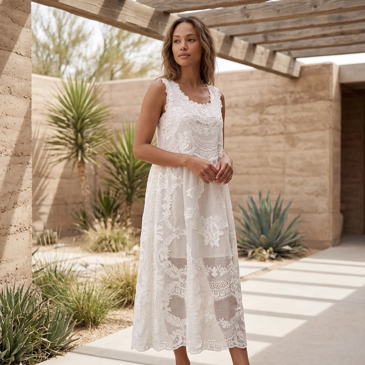 A woman models a sleeveless, pure white ethereal lace maxi dress with a scalloped hemline, standing in a sunlit outdoor architectural setting with desert plants.