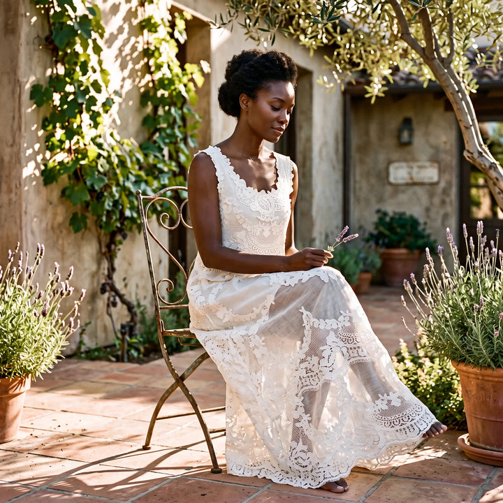 A white, sleeveless A-line floral lace maxi dress is beautifully worn by a model seated on an ornate chair in a sunlit courtyard, surrounded by greenery and lavender.