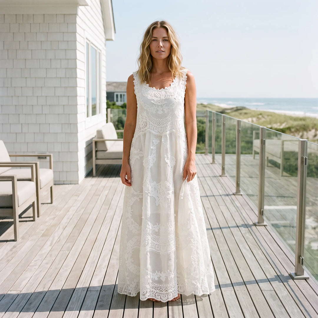 Woman wearing a sleeveless white ethereal lace A-line maxi dress with a scalloped hem, standing on a wooden deck overlooking the ocean. Romantic vacation style.