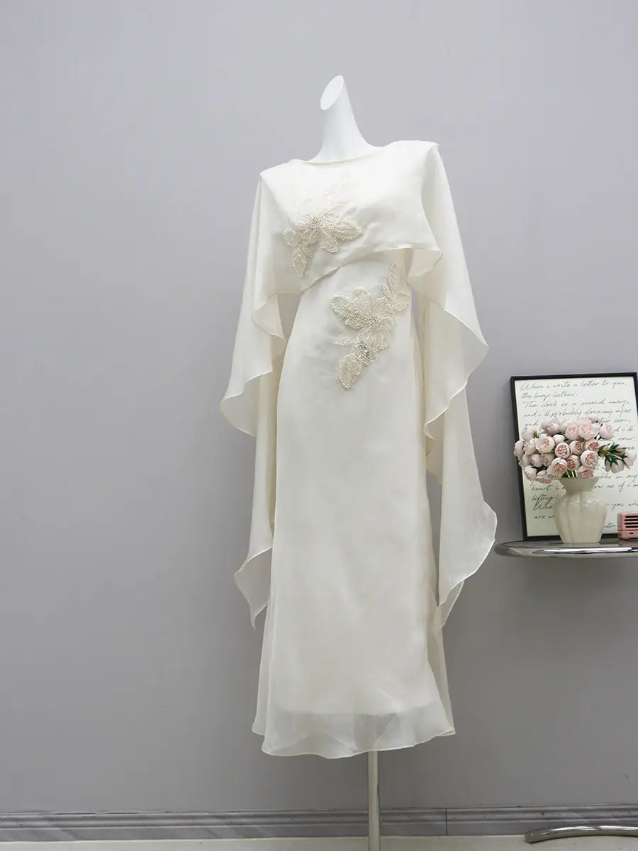 Elegant ivory formal gown on a mannequin, featuring intricate pearl floral embroidery, a boat neckline, and a flowing sheer chiffon cape. Floor-length columnar silhouette.