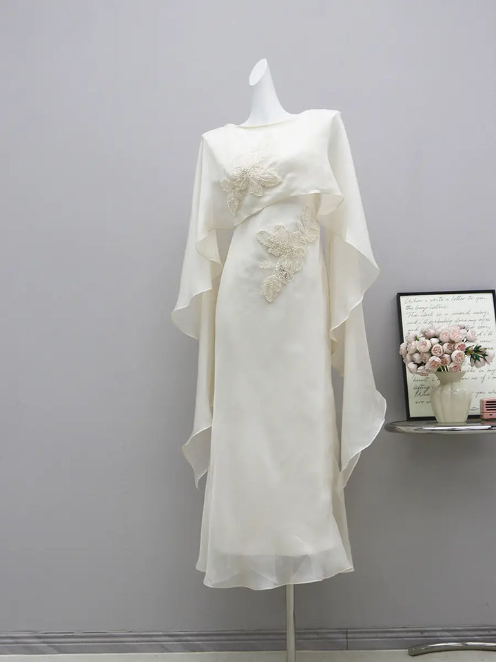 Elegant ivory formal gown on a mannequin, featuring intricate pearl floral embroidery, a boat neckline, and a flowing sheer chiffon cape. Floor-length columnar silhouette.