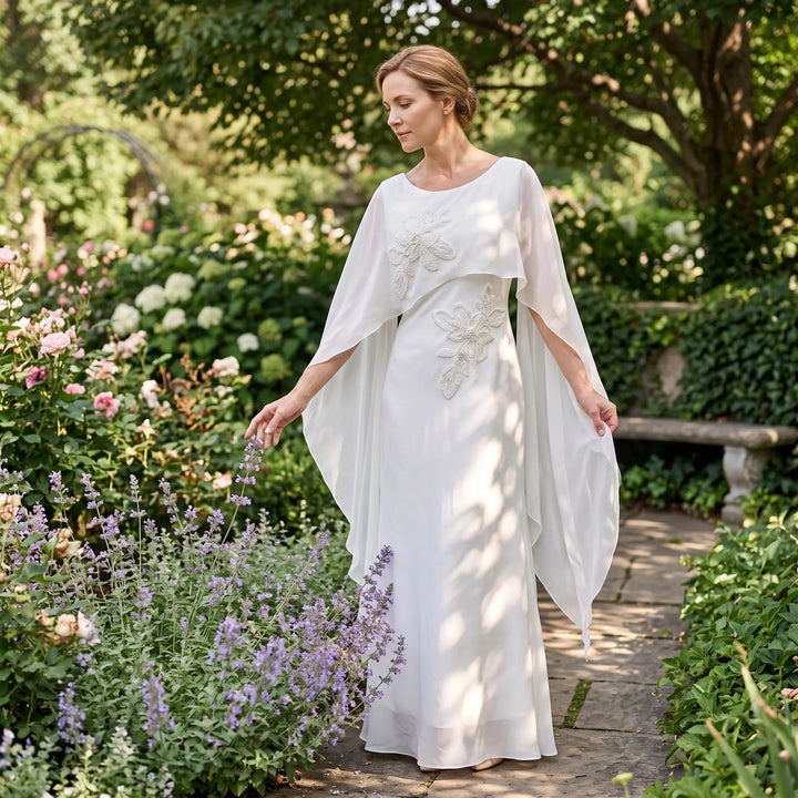 Elegant ivory satin column gown with intricate pearl floral embroidery and a sheer flowing chiffon cape, worn by a woman in a lush garden setting.