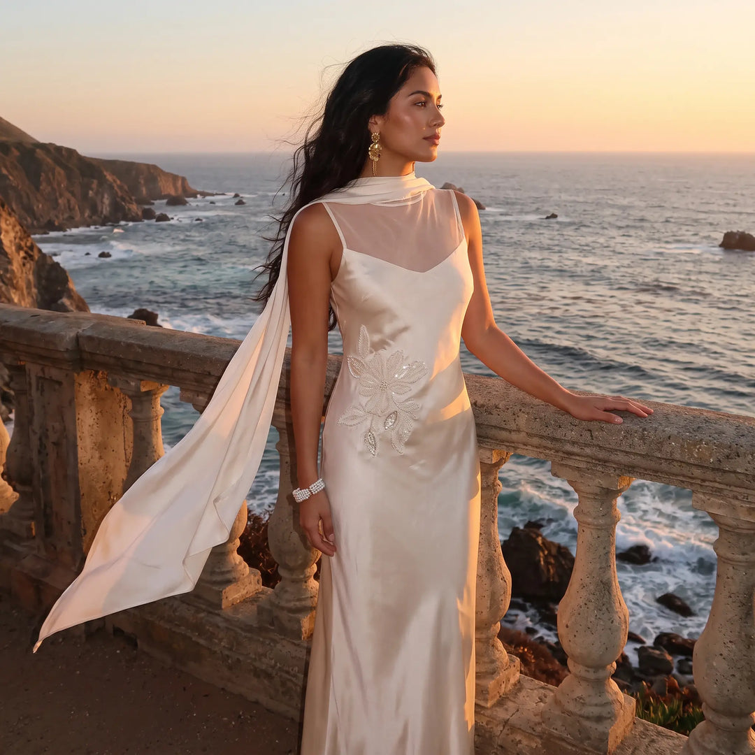 An elegant ivory satin Zaylin formal gown with pearl embroidery and a flowing sheer chiffon cape, modeled by a woman standing on a stone balcony overlooking the ocean at sunset.