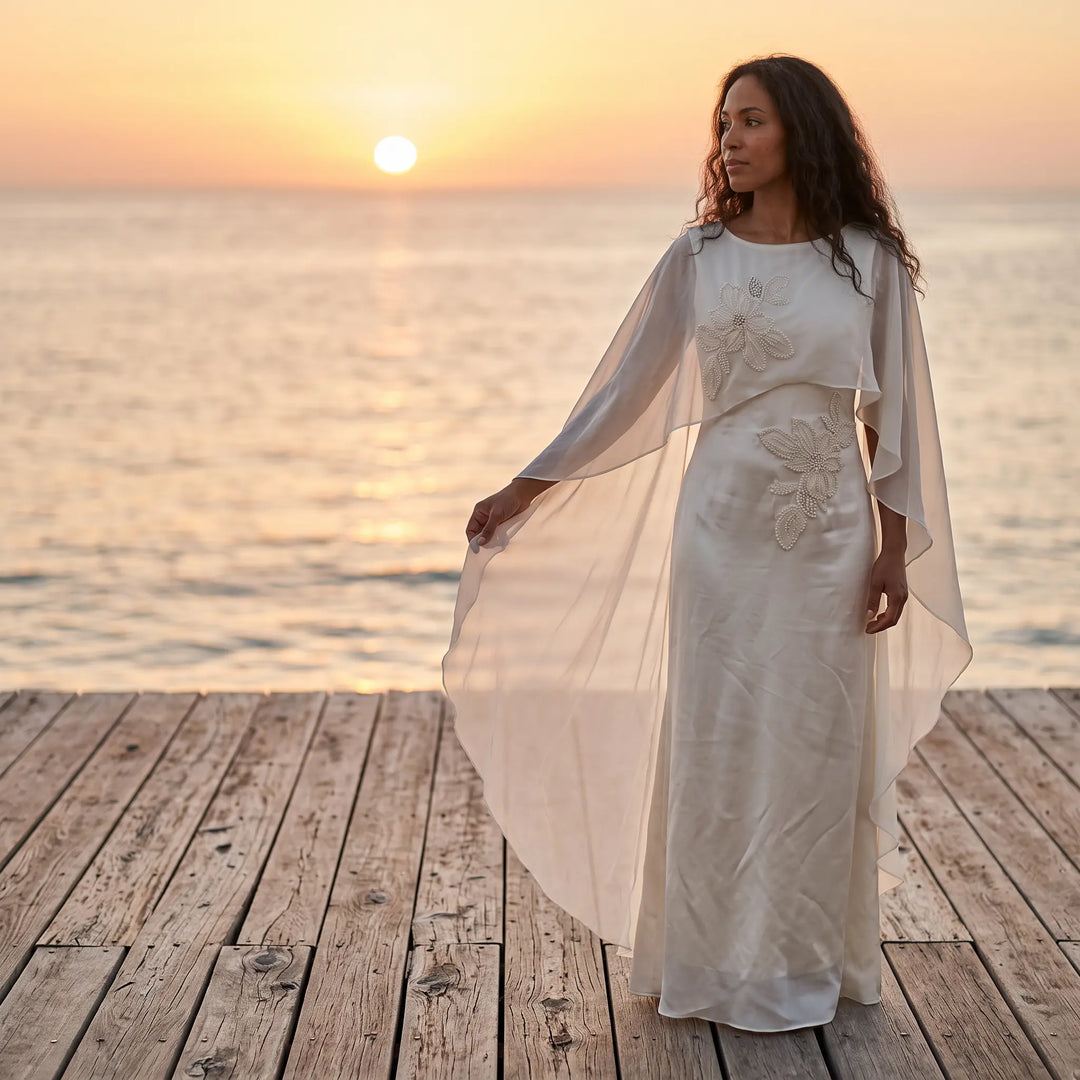 Elegant ivory satin column gown with intricate pearl floral embroidery and a flowing sheer chiffon cape, worn by a model standing on a wooden dock at sunset.